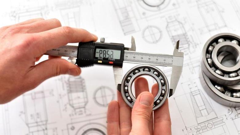 A quality engineer measuring a ball bearing with calipers to ensure its compliance, with a reference diagram in the background.