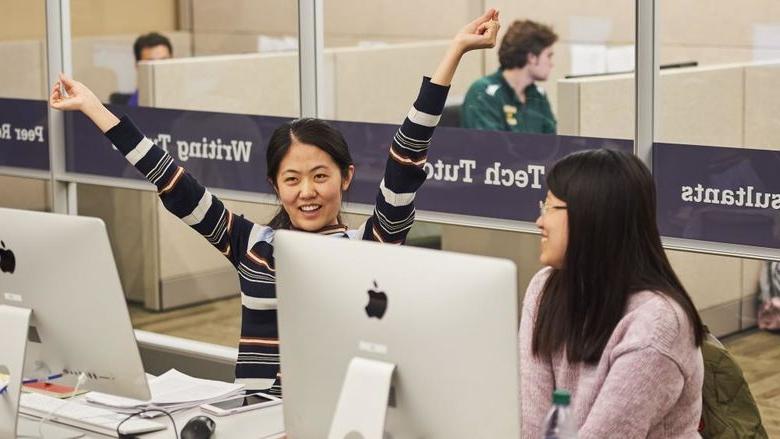 two students sitting together at computer stations, student at right smiles with arms raised joyfully